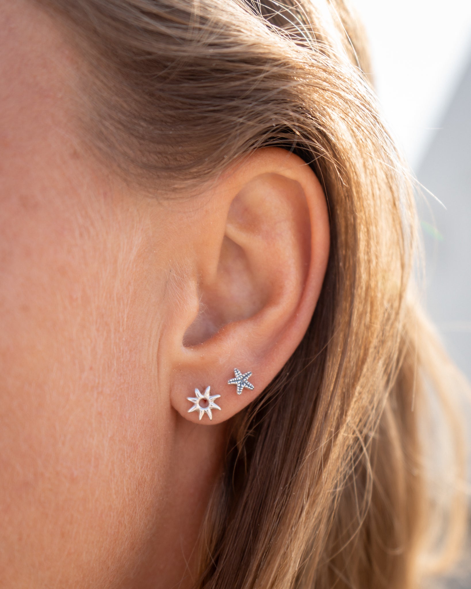 Silver star and sun stud earrings worn on ear in natural sunlight, ocean-proof jewelry.