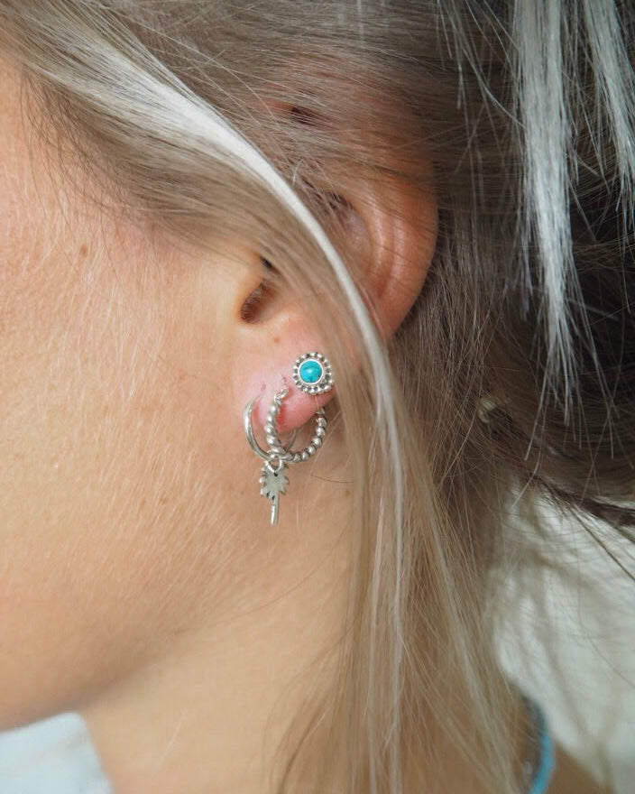 Turquoise stud earring worn in a stacked ear look with silver hoops and charm details in natural light